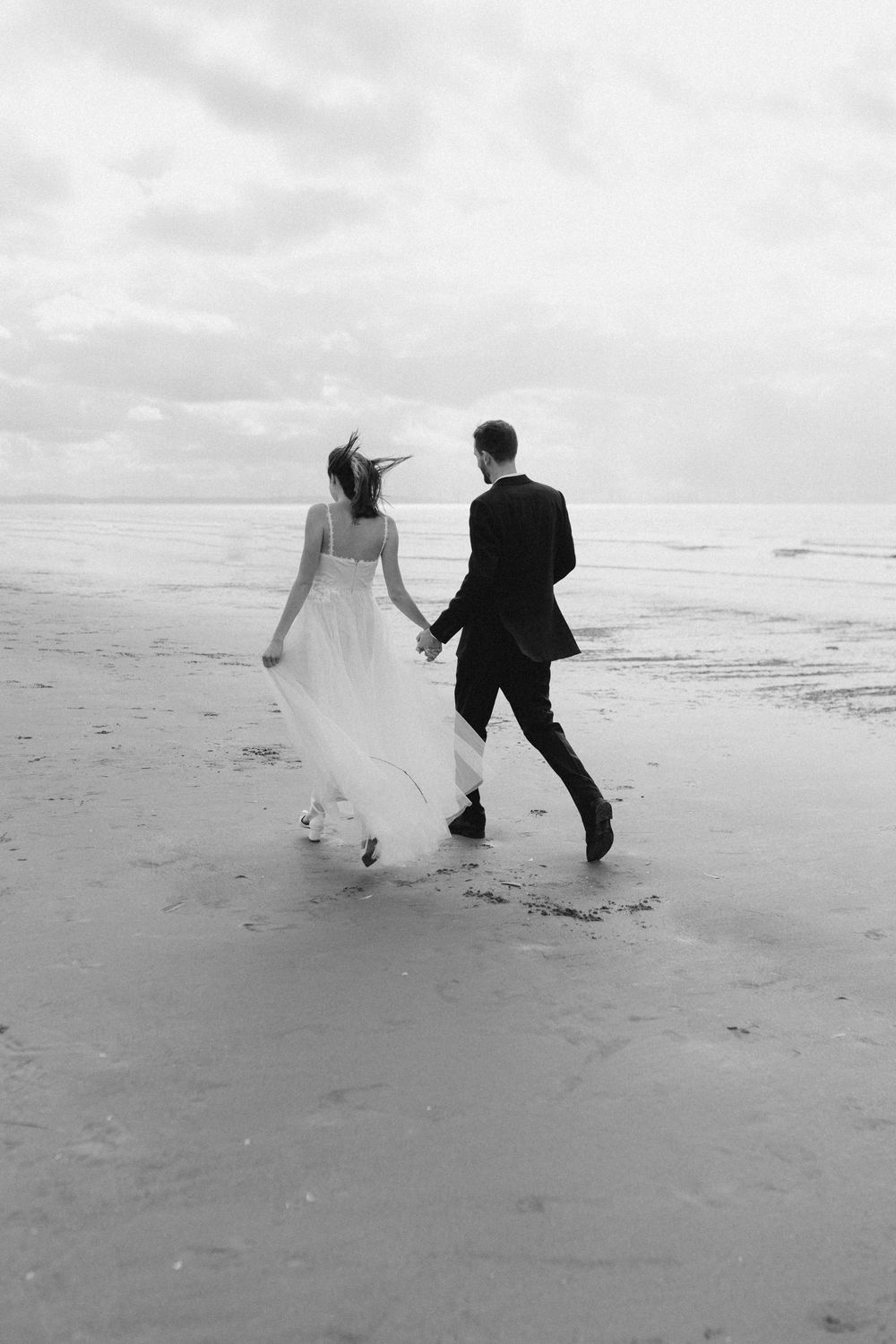 A couple in wedding attire runs playfully along a misty beach shoreline in black and white.