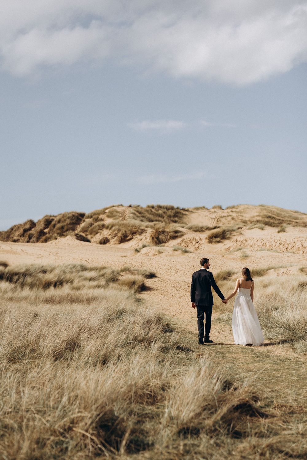 A couple in wedding attire walks hand in hand through golden beach grass on sand dunes under cloudy skies.