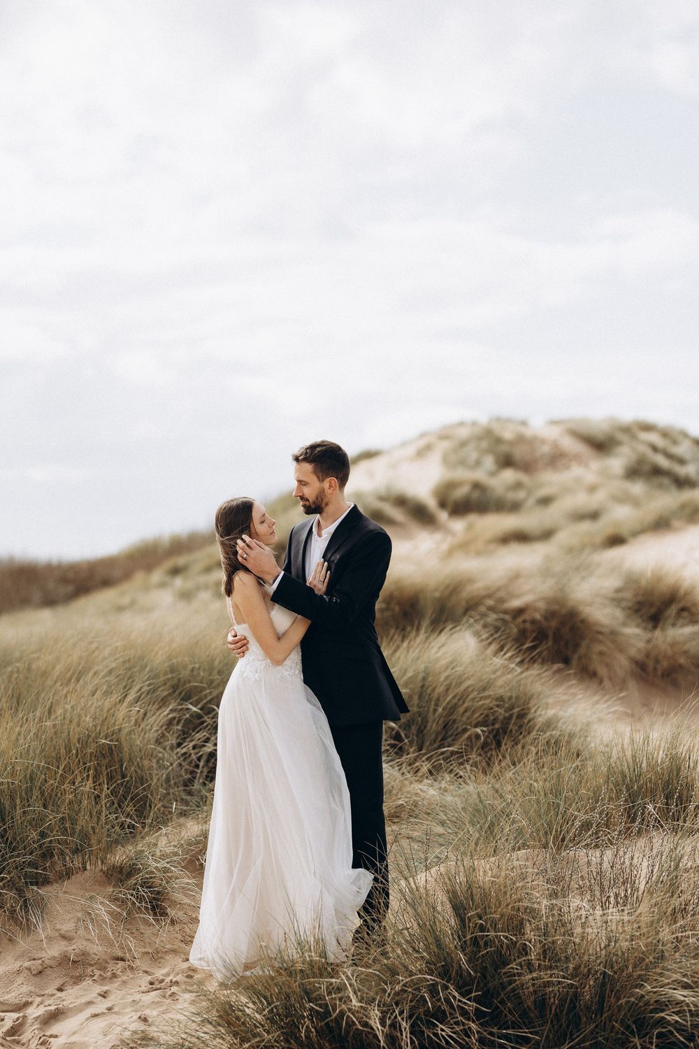 A romantic wedding couple embraces in tall beach grass against a cloudy sky.