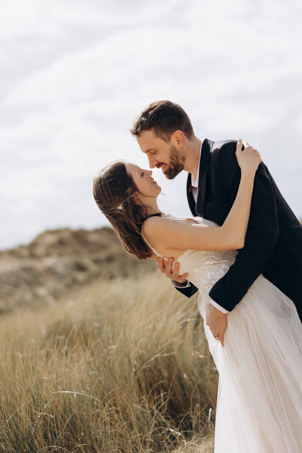 A couple shares an intimate dance pose amid windswept coastal dunes.