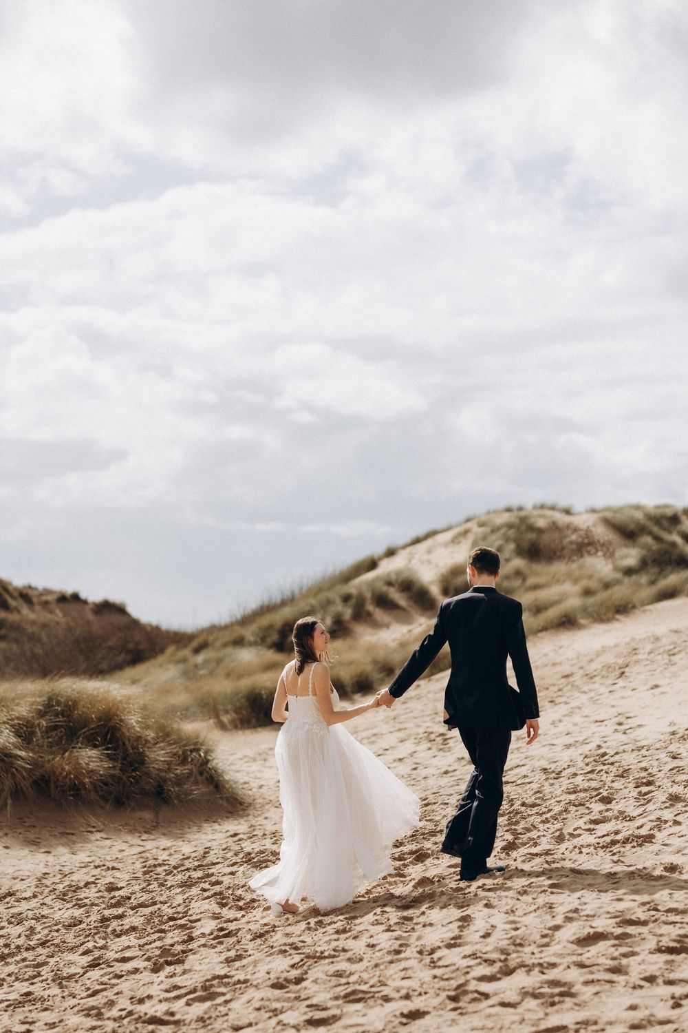 A couple in wedding attire walks up sandy dunes together under cloudy skies in a romantic beach setting.