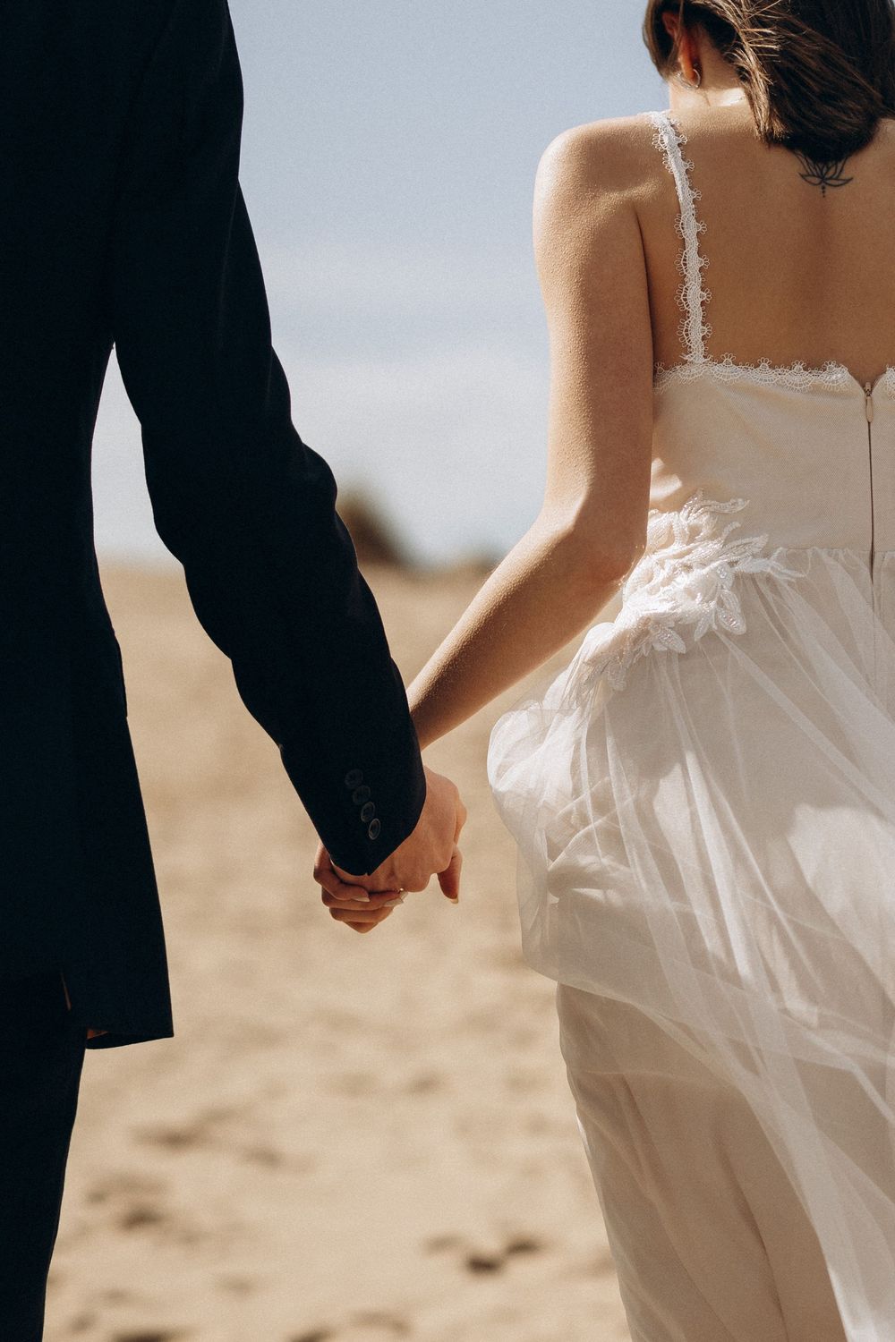 Couple holding hands on sandy beach with bride wearing flowing white wedding dress in romantic beach wedding scene.