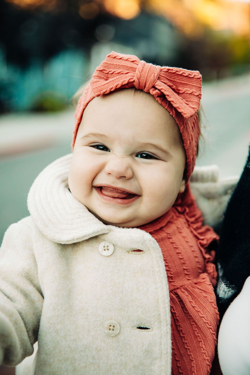 Baby wearing cream colored coat and coral pink bow headband smiles joyfully outdoors.