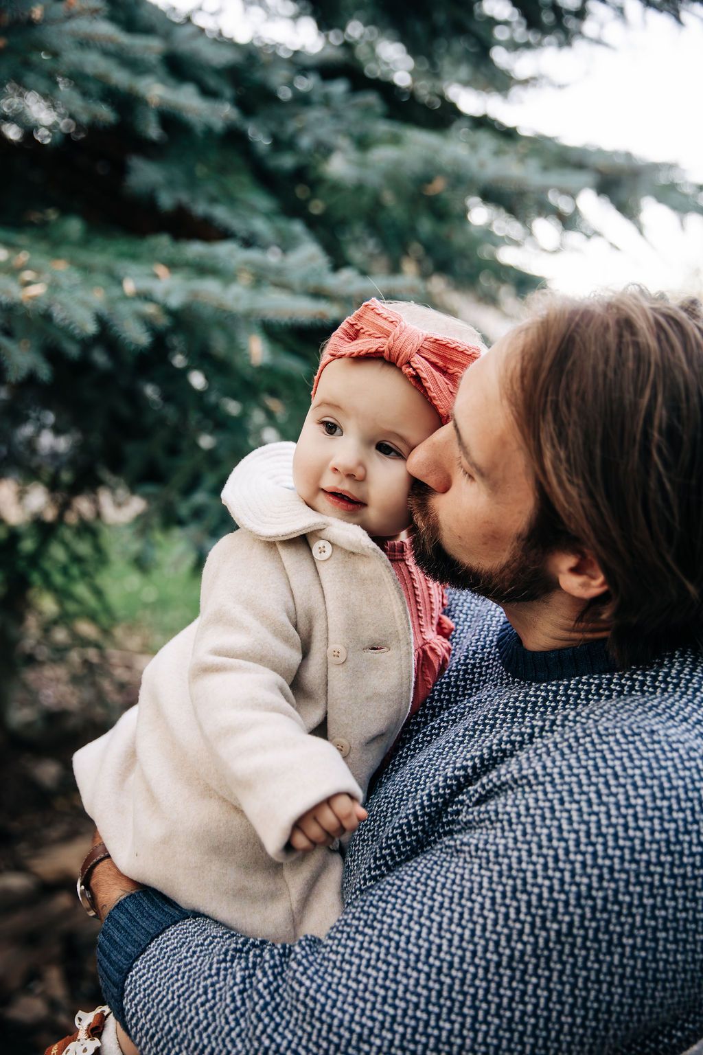 A tender moment between parent and baby in a cream coat with a red bow headband against an evergreen backdrop.