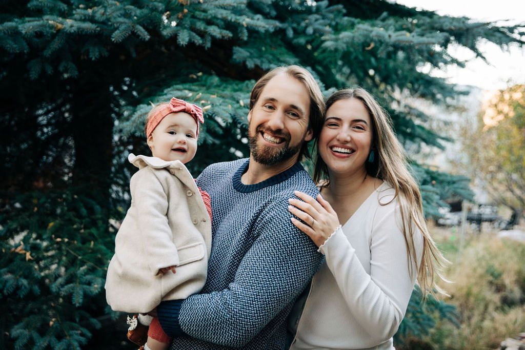 A joyful family laughs and embraces during an outdoor photo session against a backdrop of evergreen trees.