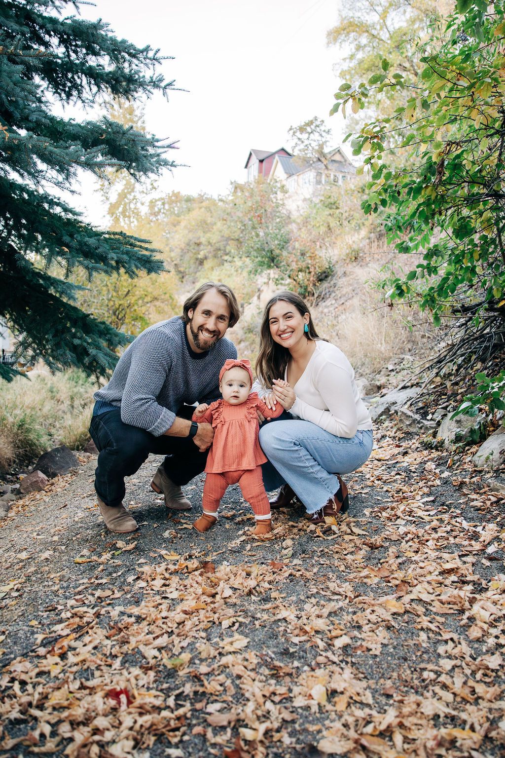 A family enjoying autumn outdoors together on a leaf-covered path surrounded by evergreen trees.