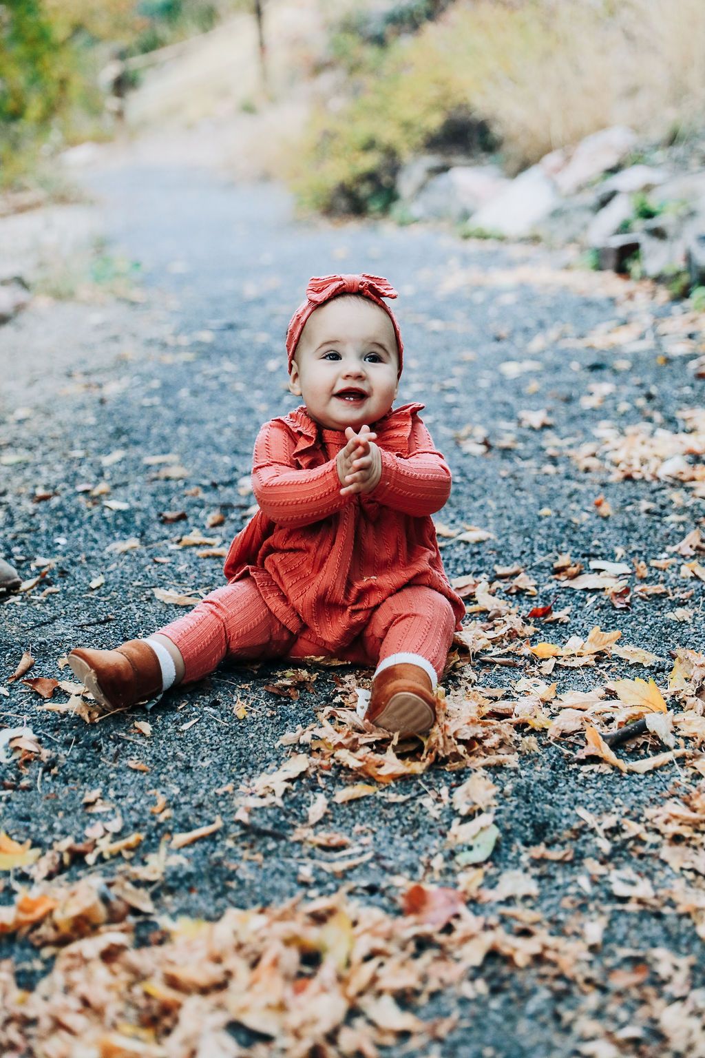 Small child in red outfit sits among autumn leaves on ground during fall photo session.