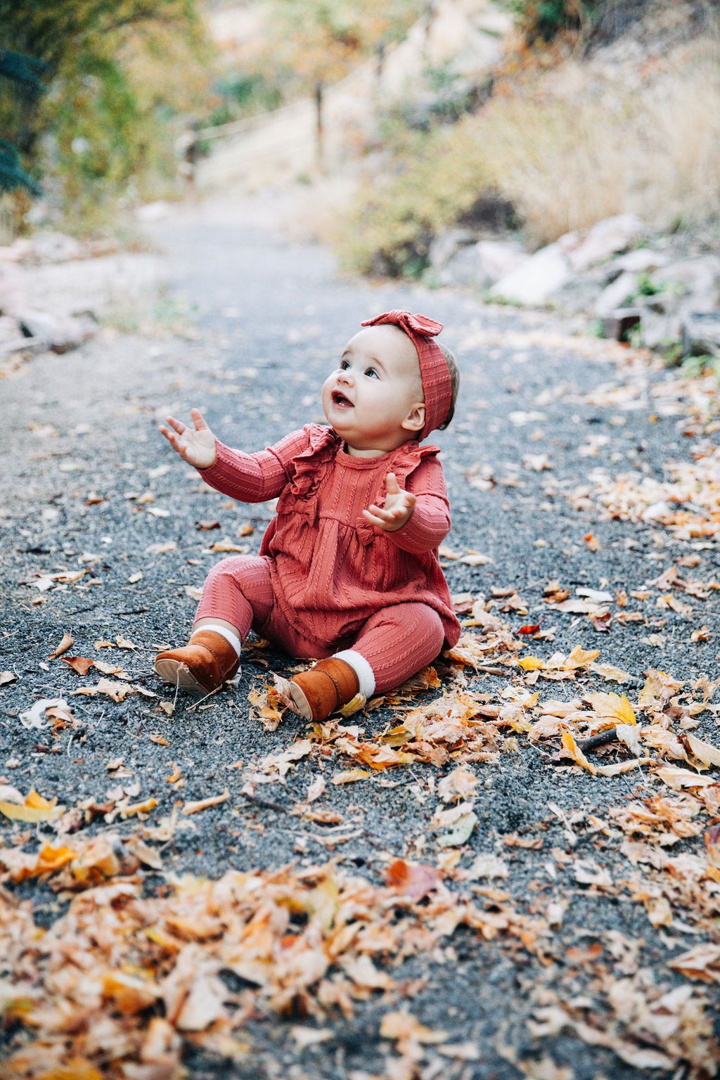 Small child wearing burgundy sweater plays joyfully in autumn leaves scattered along a forest path.