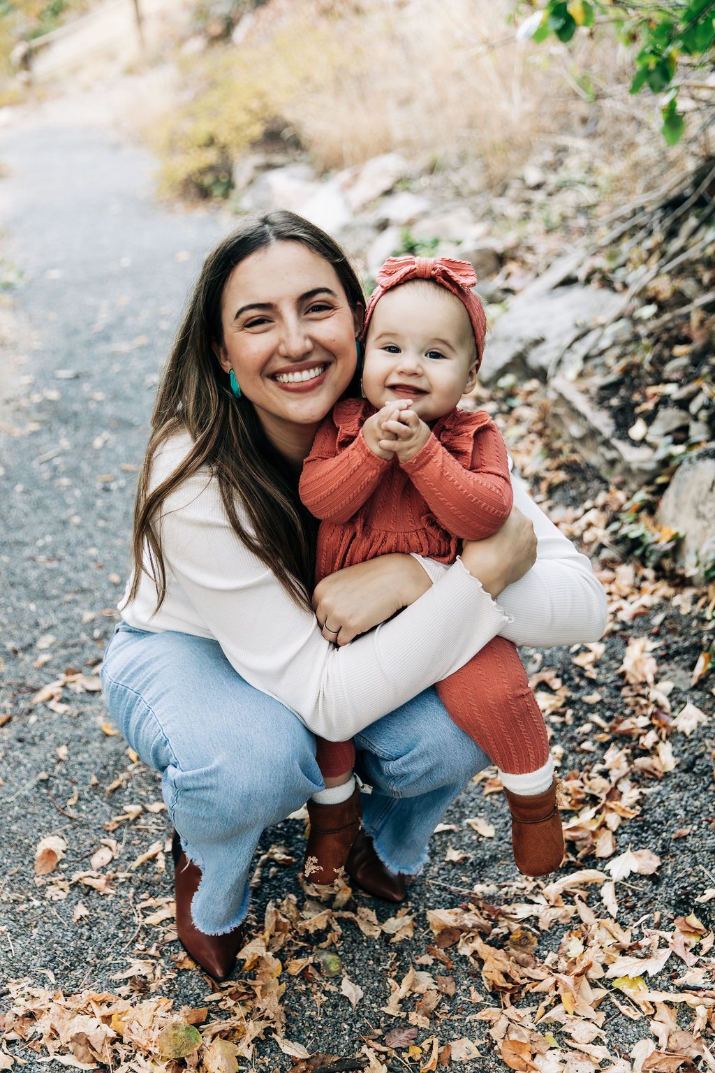 A heartwarming autumn scene of parent and baby in matching coral outfits sitting on a leaf-covered path.