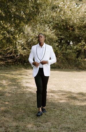 Groom in white tuxedo jacket stands alone in a sunny outdoor setting.