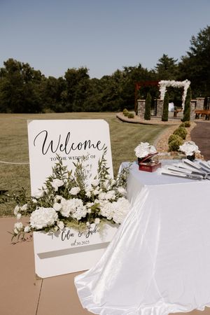 White wedding welcome sign and floral decorations set up on outdoor tables.