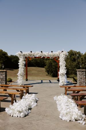 White floral arrangements decorating wooden arbor with stone pillars at outdoor venue.
