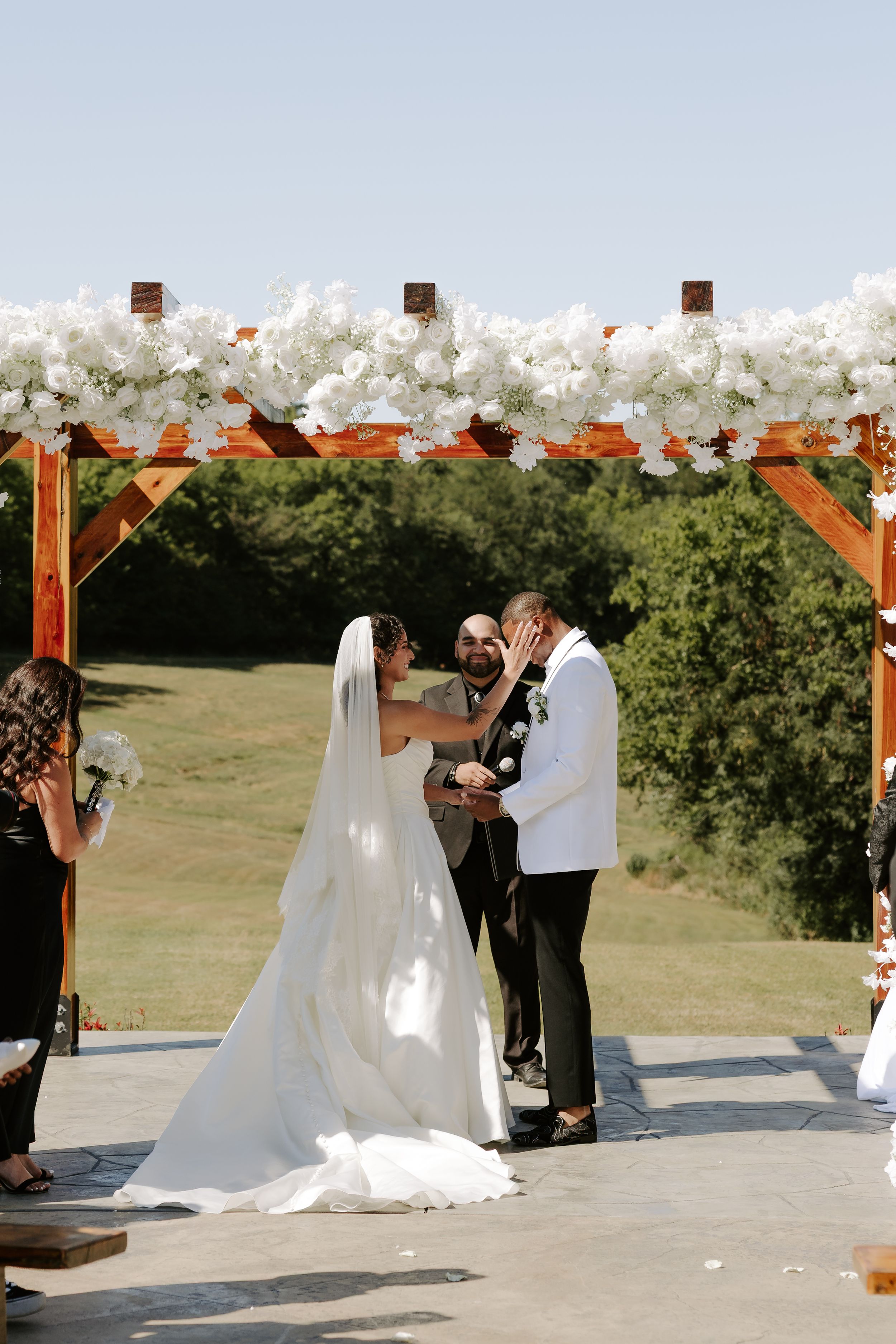 An outdoor wedding ceremony under a wooden arch decorated with white floral arrangements.