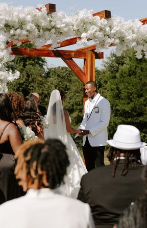 Wedding ceremony under a wooden arch decorated with white flowers and flowing fabric.