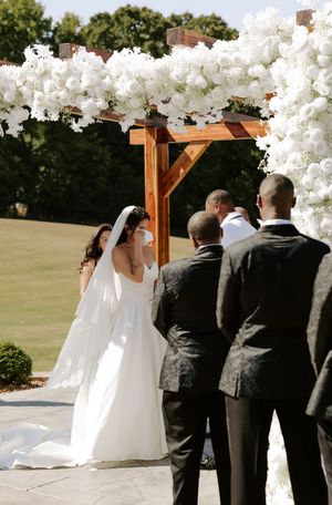 Wedding party gathers under a floral-decorated wooden arch during outdoor ceremony.