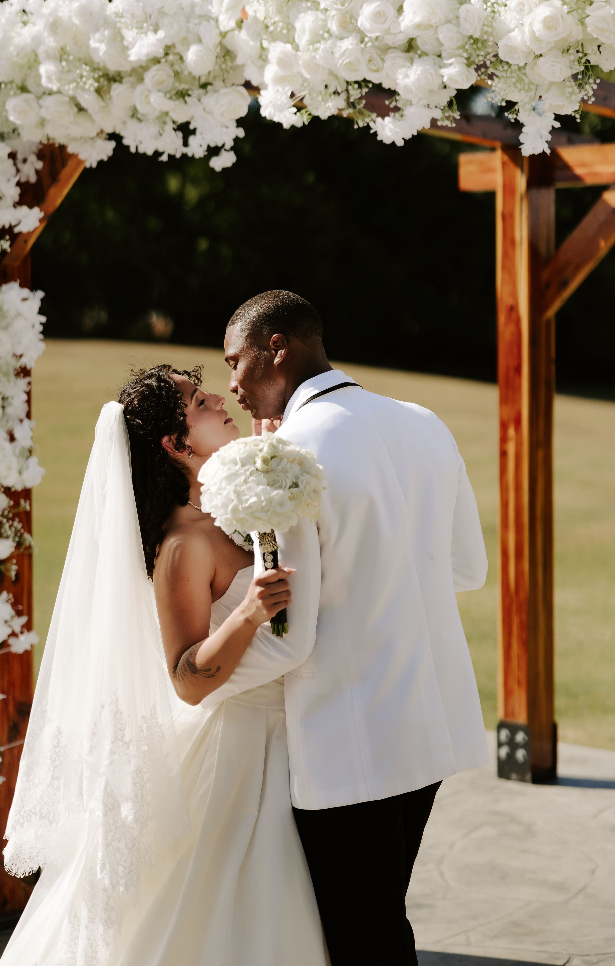A bride and groom share their first kiss under a floral-decorated wooden arch at an outdoor wedding.