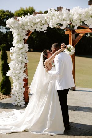 A couple embraces under a white floral arch during their outdoor wedding ceremony.