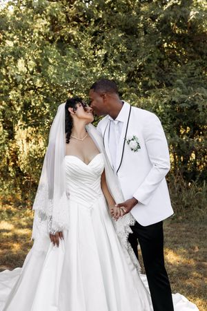 A romantic wedding portrait of a couple in white attire embracing outdoors with sunlit trees in the background.