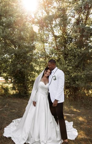 A romantic wedding photo of a couple in white attire with sunlight streaming through trees.