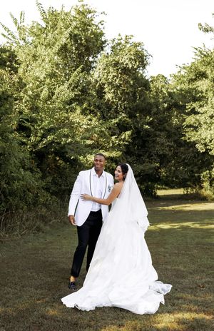 An elegant outdoor wedding portrait featuring a flowing white dress and classic white tuxedo jacket.