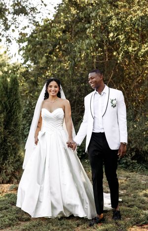 Wedding couple in white attire walking together through a garden setting at sunset.