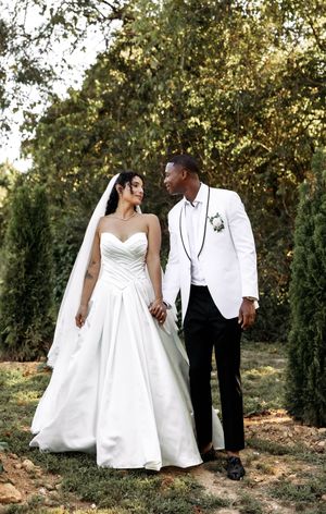 Joyful wedding couple poses in white formal attire against a natural outdoor backdrop.