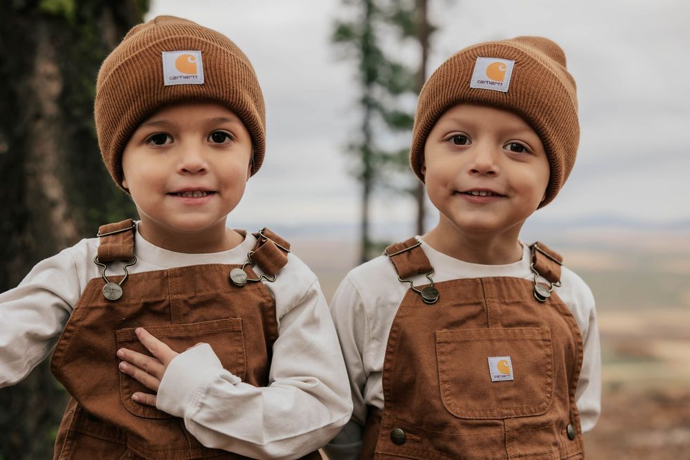 Two kids in matching brown Carhartt overalls and beanies pose together.