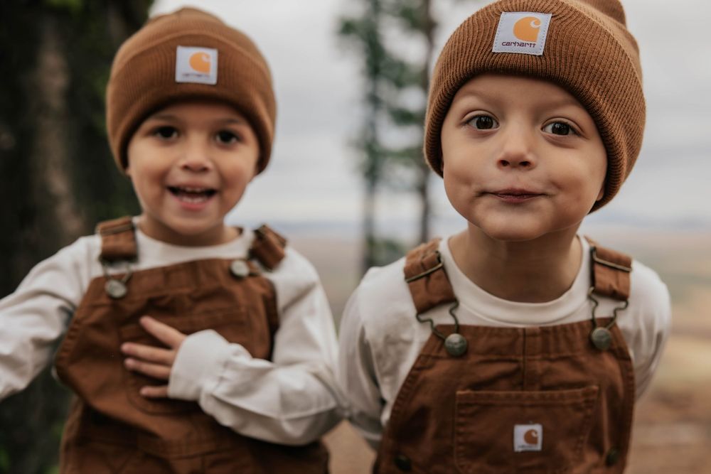 Young children wearing brown Carhartt beanies and overalls smile at camera.