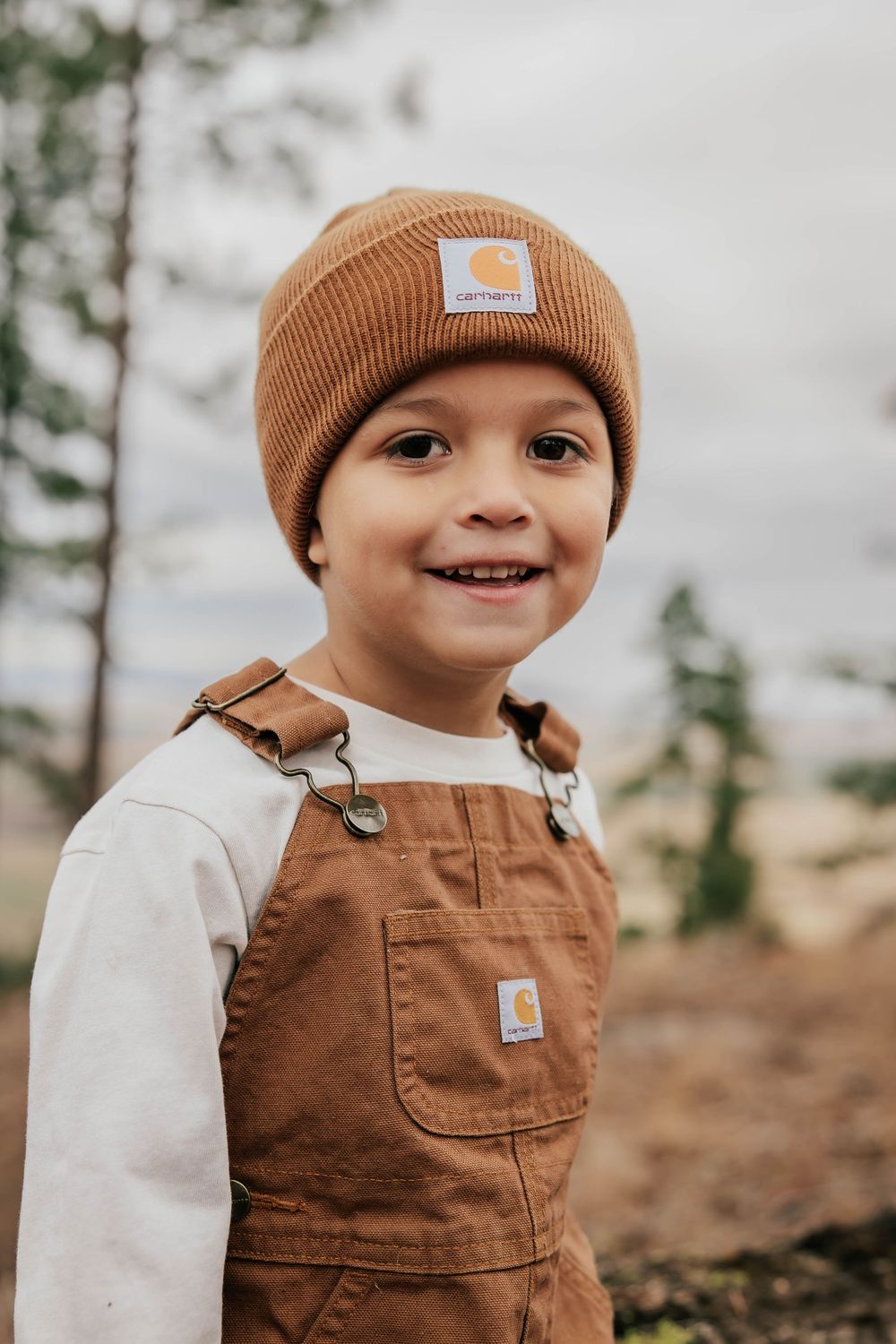Child in brown Carhartt beanie and overalls smiles outdoors.