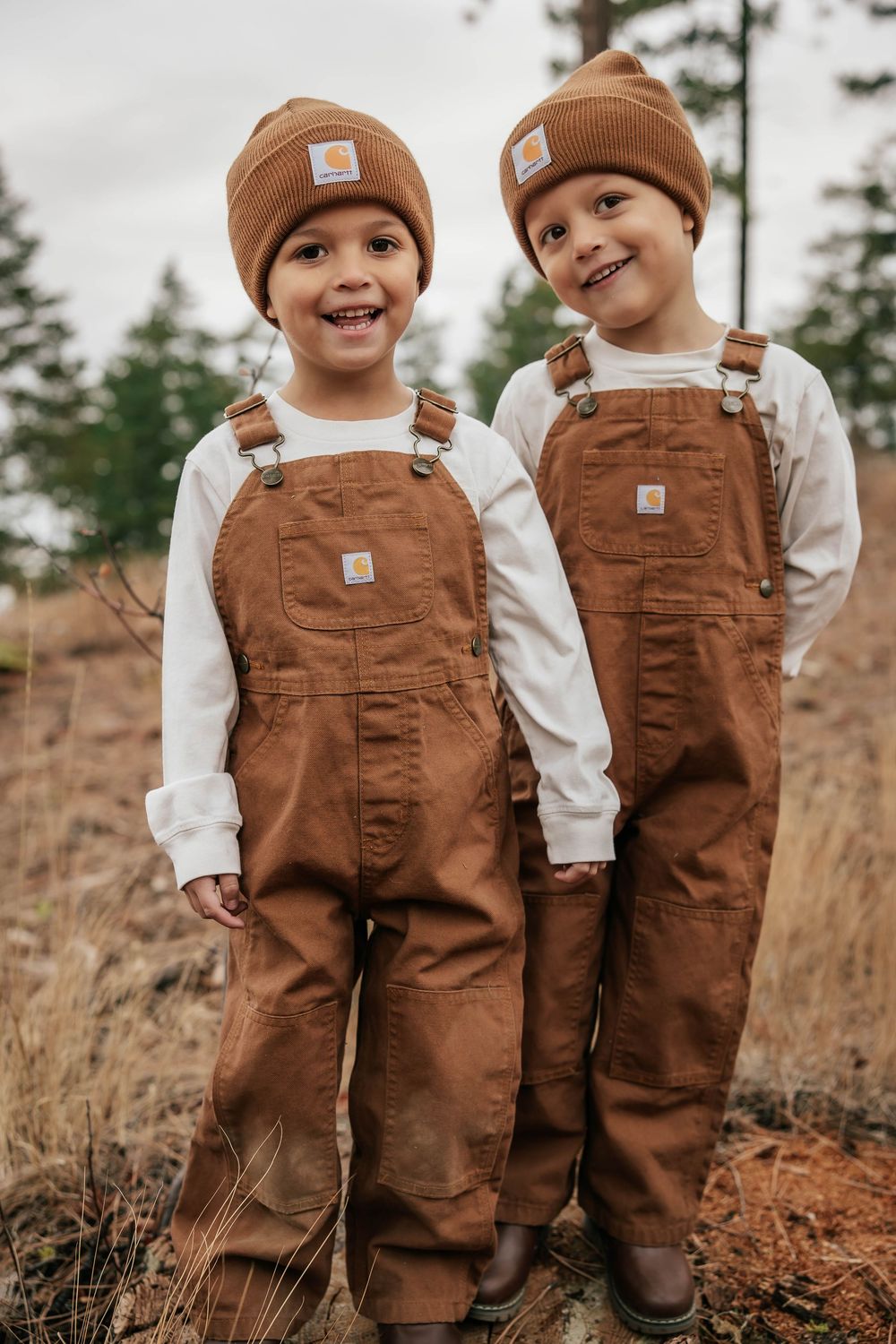 Two children in matching brown overalls and beanies laugh together on hillside.
