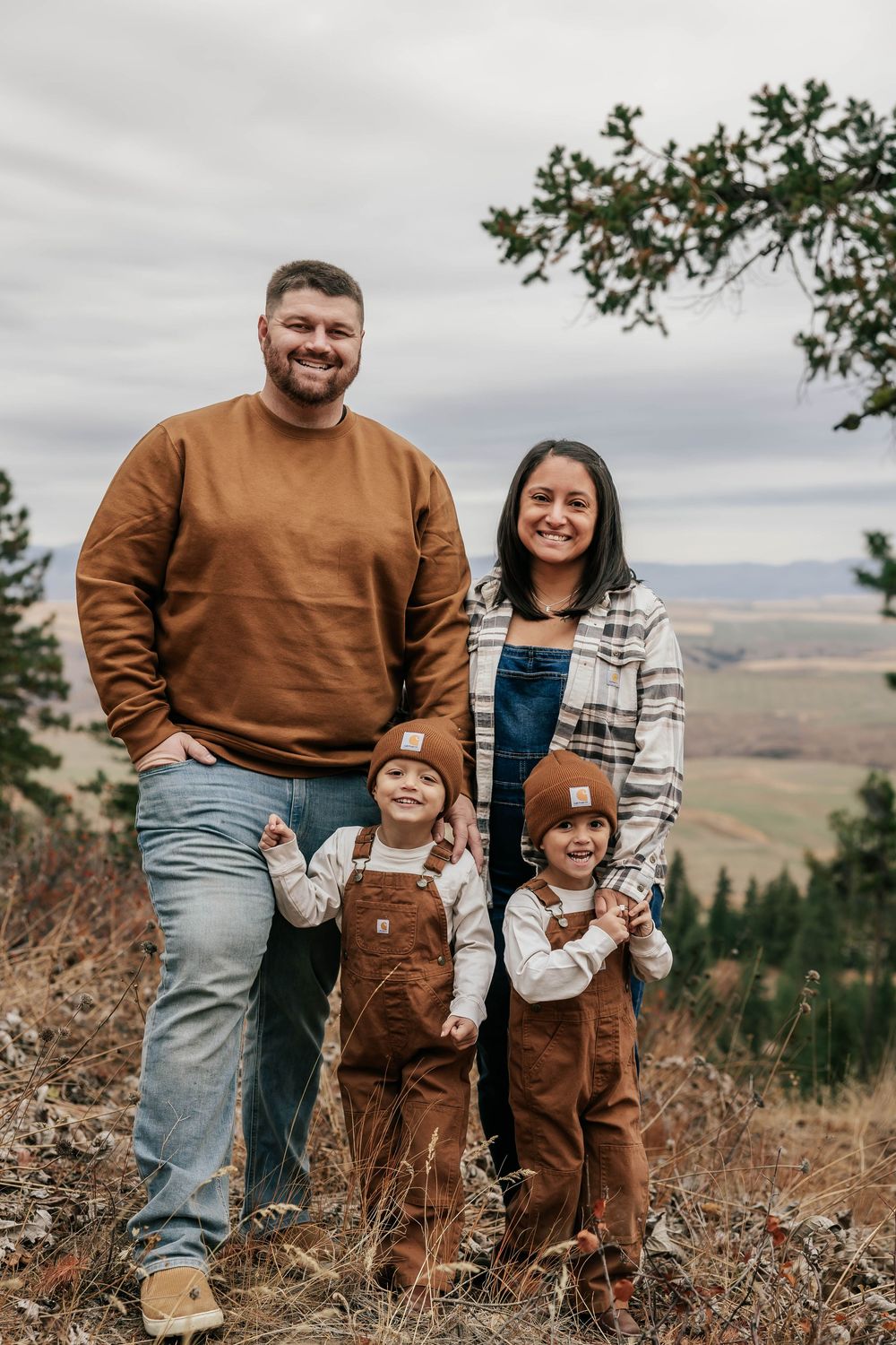A family in coordinated brown outfits poses together on a hillside with mountains in the background.