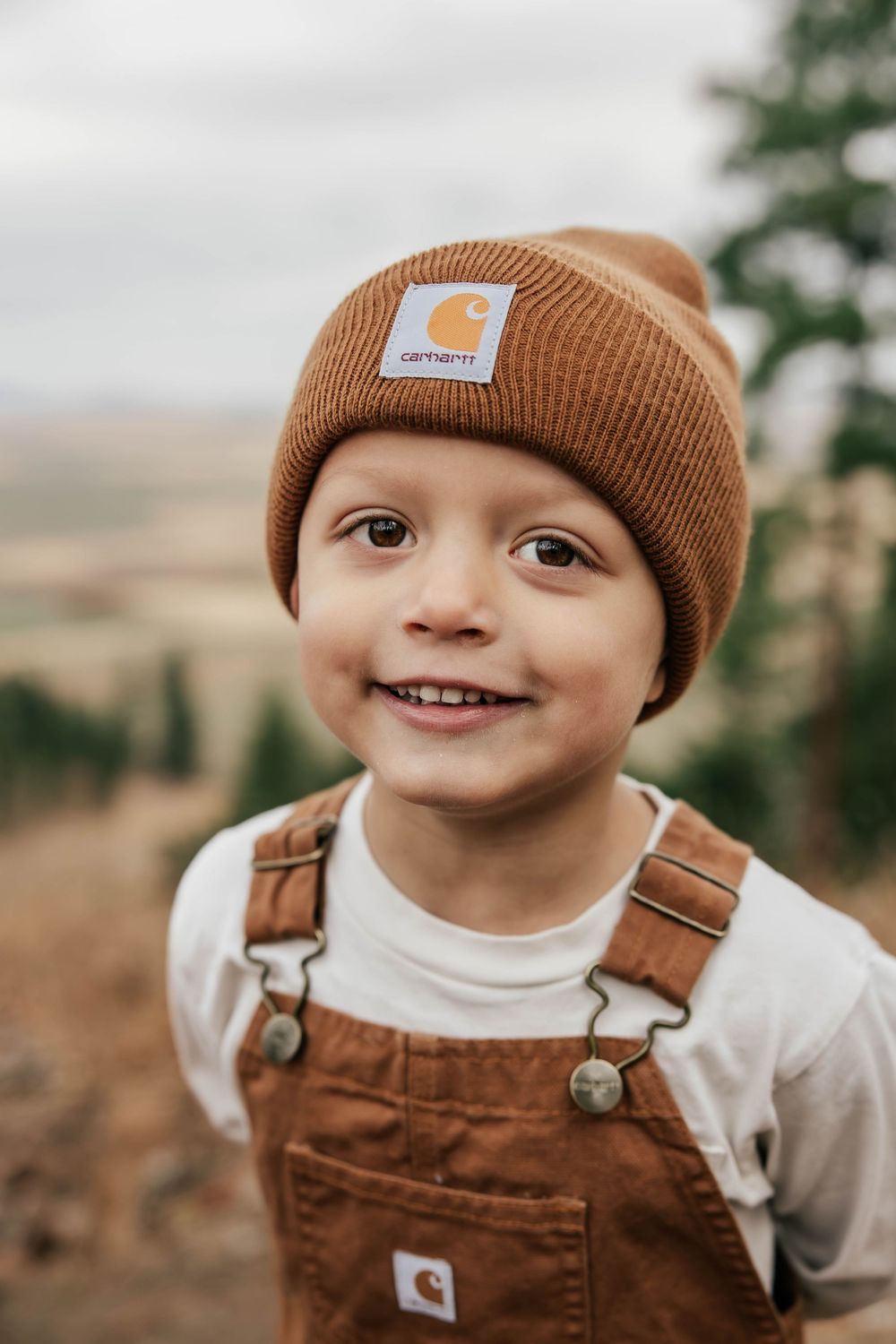 Close up of a small child wearing brown overalls and a brown beanie hat smiling at the camera.