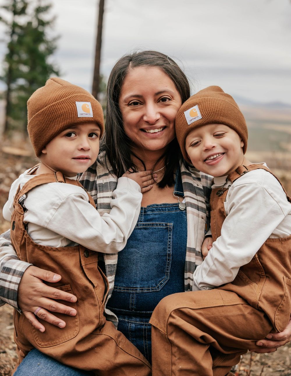 Three people wearing matching brown Carhartt beanies and overalls pose together outdoors against a cloudy landscape.