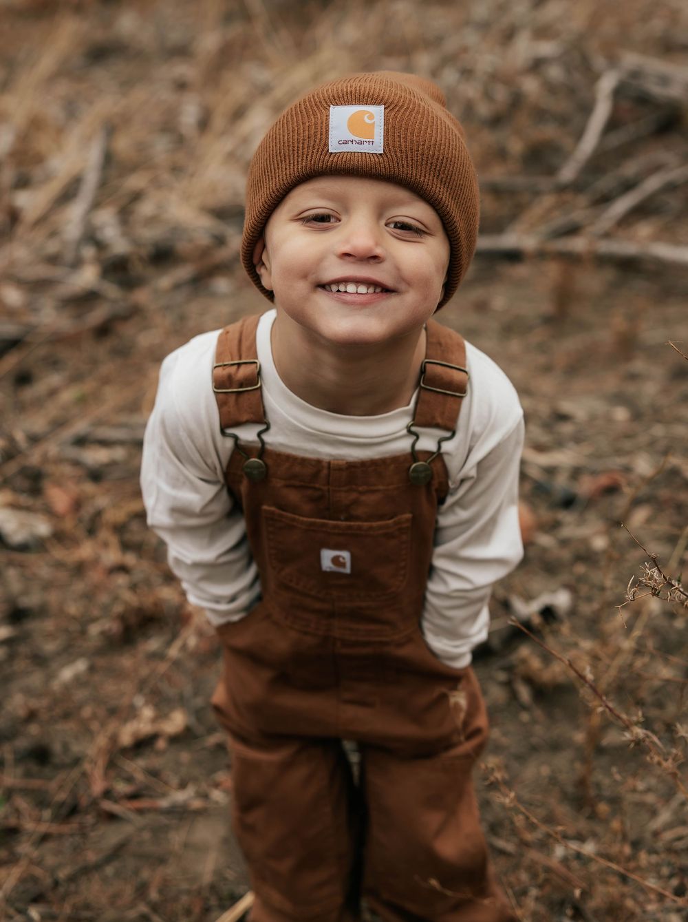 Child in brown overalls and matching beanie poses cheerfully outdoors.