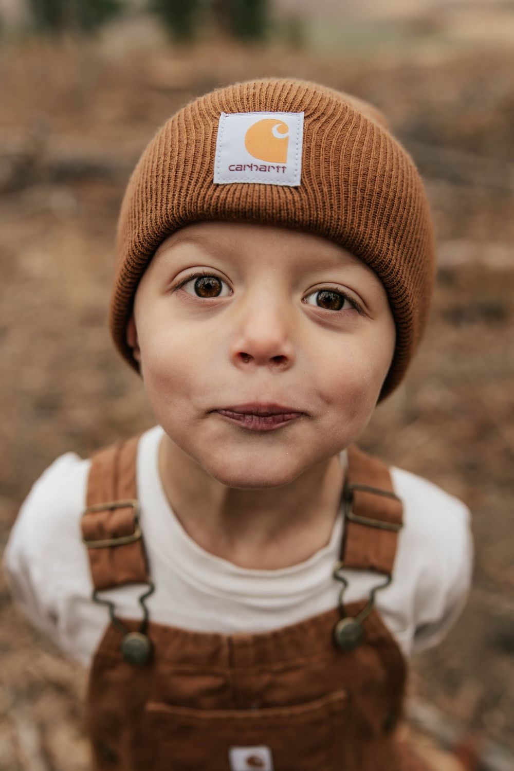 A close-up portrait of a child wearing a brown Carhartt beanie and tan overalls.