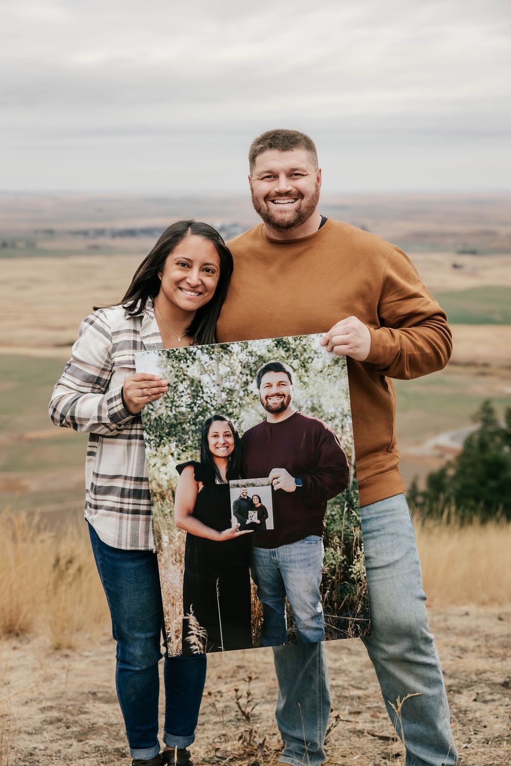 A couple stands outdoors holding photos in a photo-inception style against rolling hills.