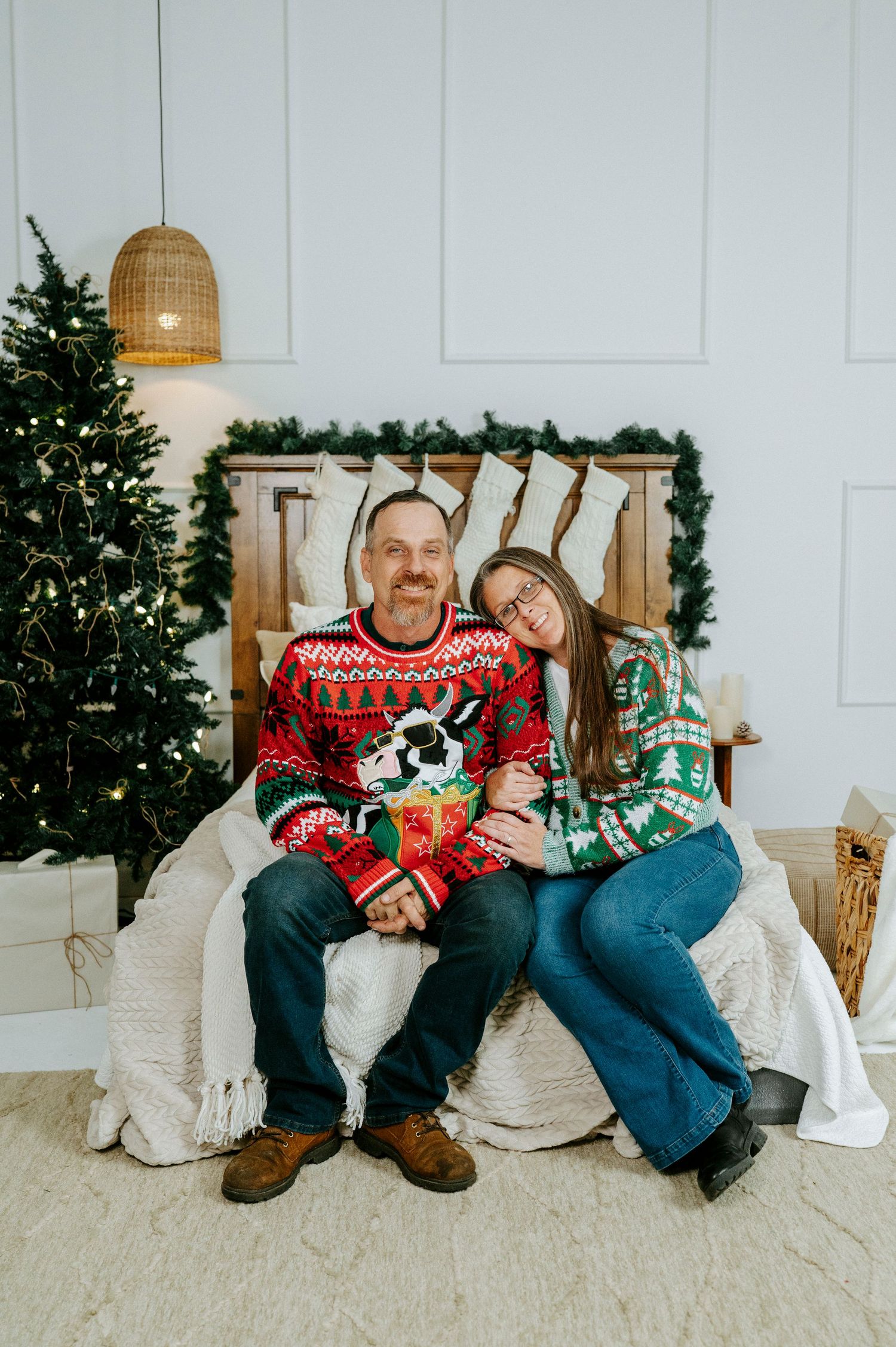A couple in festive Christmas sweaters sits together on a white couch with a decorated tree in the background.