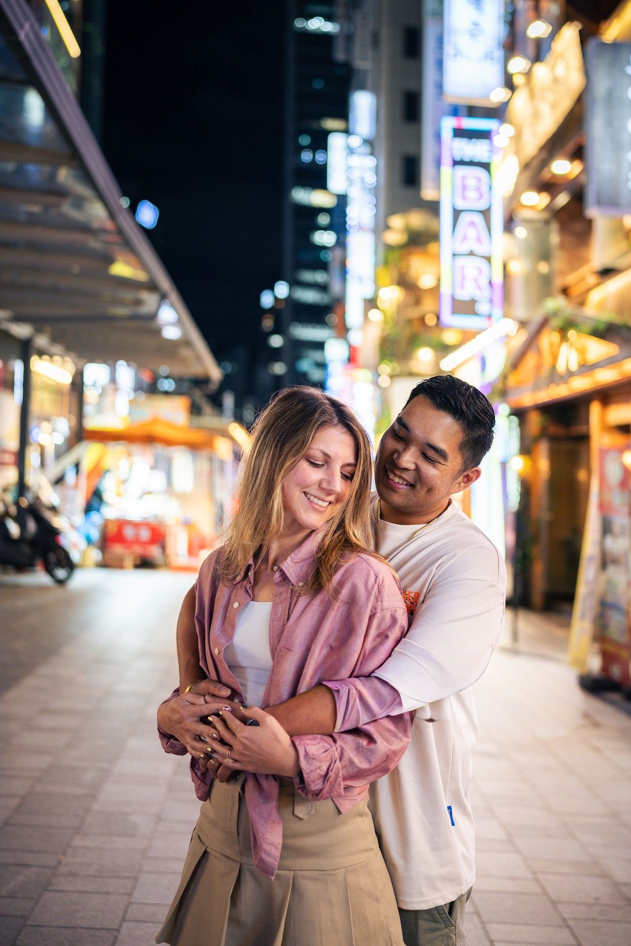 A couple embraces on a vibrant nighttime street with glowing neon signs and city lights in the background.