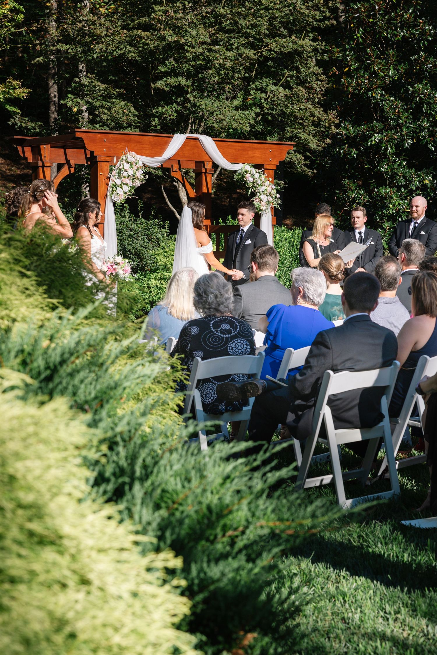 A wedding party poses outdoors on a sunny day with trees and greenery in the background.