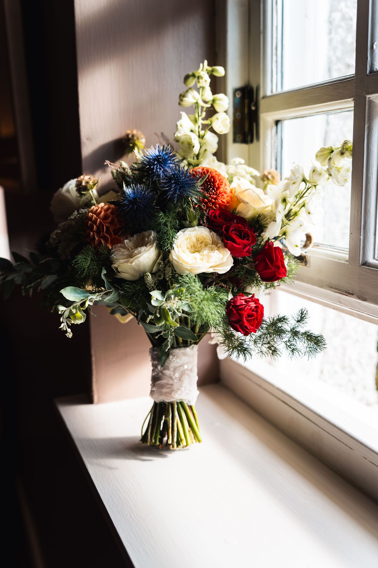 A vibrant winter bridal bouquet with red roses, blue thistles, and white flowers rests on a windowsill.