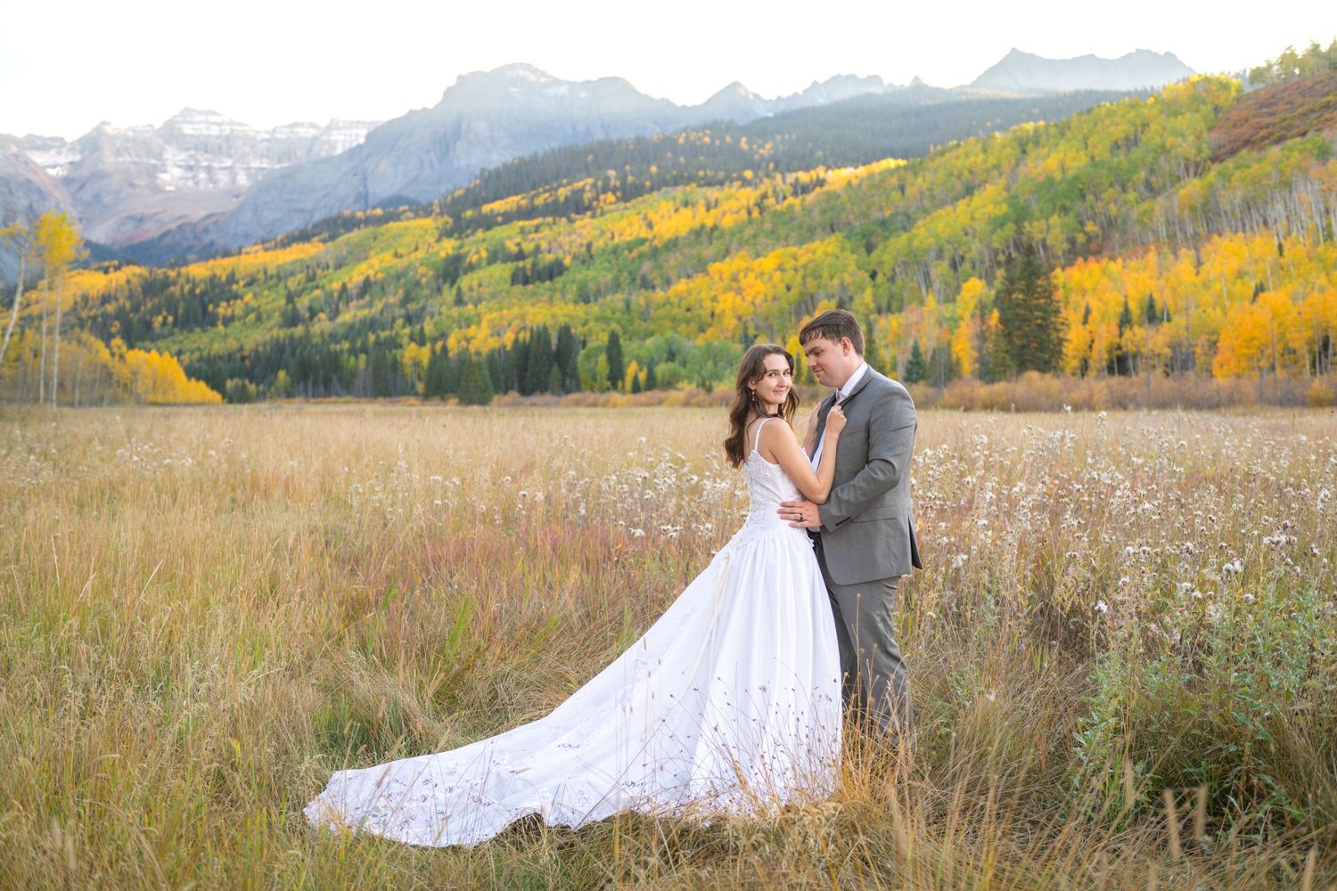 A bride and groom embrace in a golden autumn meadow with aspen trees and mountain peaks in the San Juan Mountains.