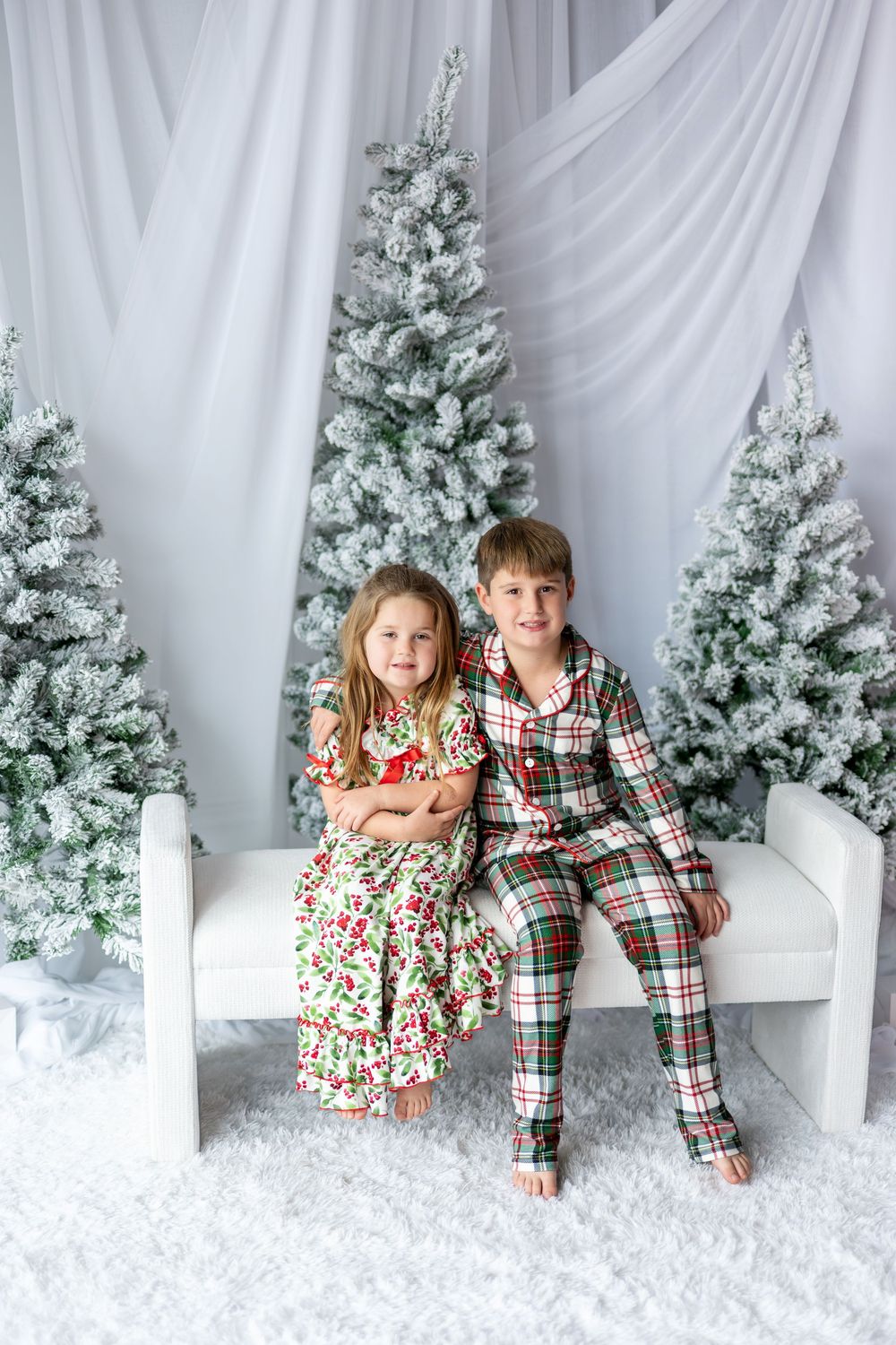Two young siblings in Christmas pajamas sit together on a white bench among snow-flocked holiday trees.
