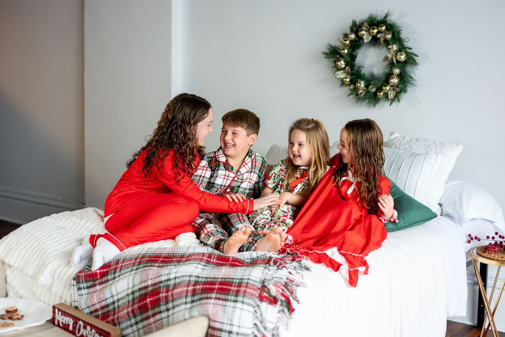 Four children in festive Christmas pajamas laugh and chat together on a cozy holiday-decorated bed.