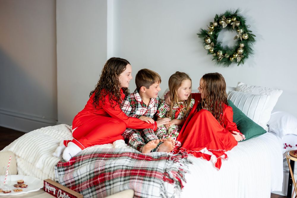 Four children wearing Christmas pajamas sit together on a bed conversing beneath a decorative holiday wreath.