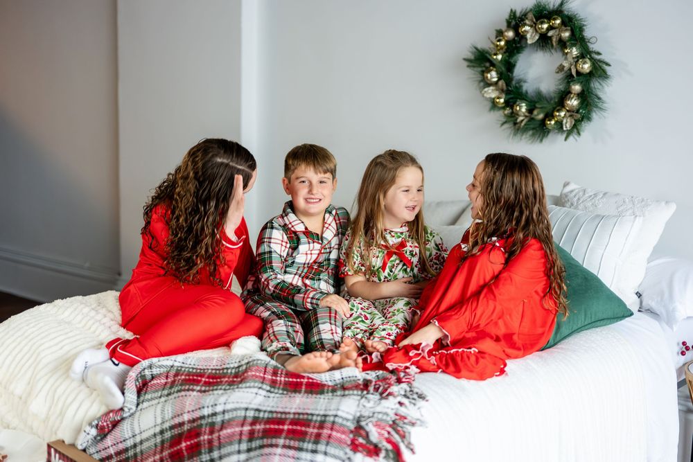 Four kids in Christmas pajamas chatting and laughing together on a cozy white bed.