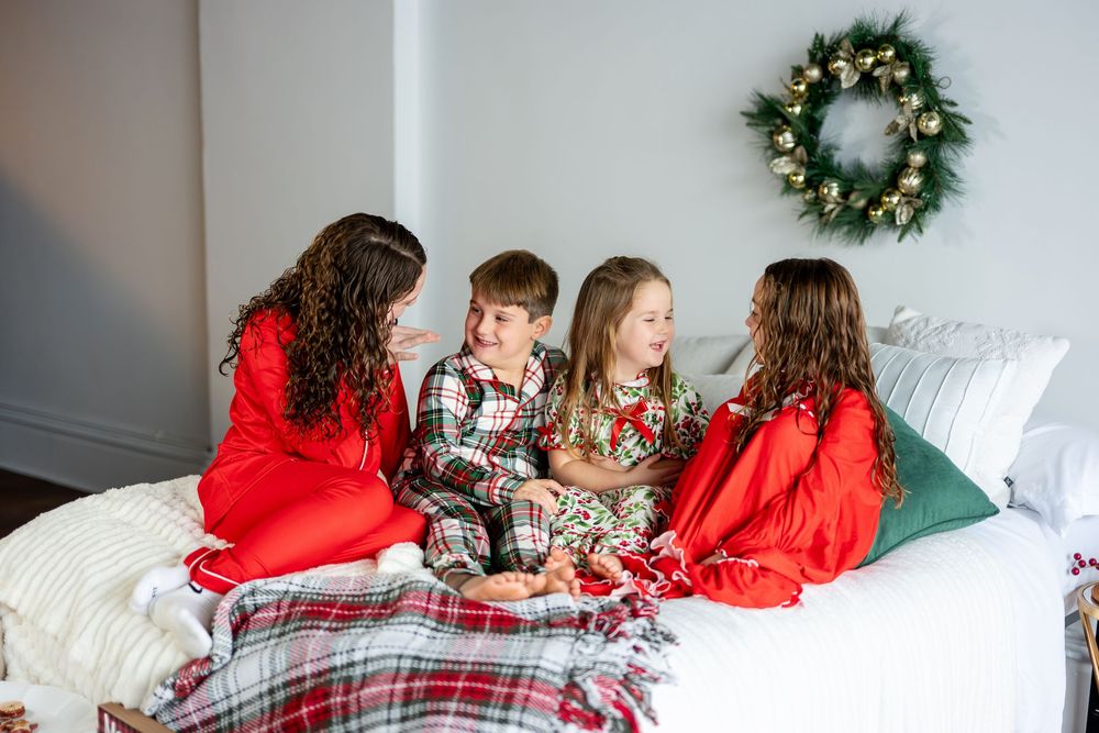 Family in Christmas pajamas laughing together on a white bed with holiday wreath decor.