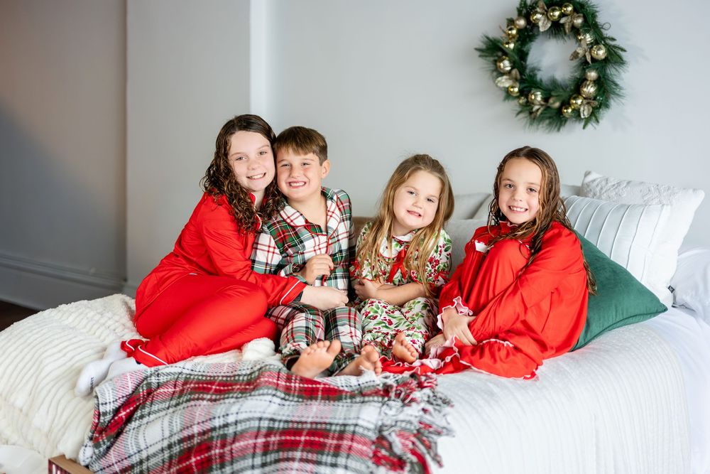 Four children in festive red and plaid Christmas pajamas sitting together on a white bed.