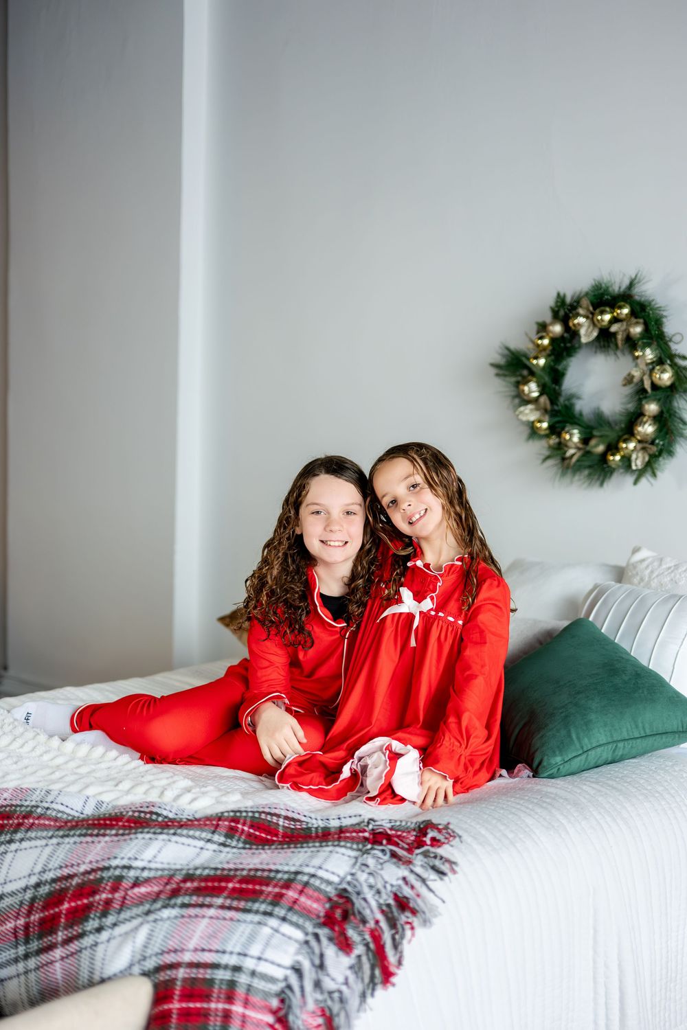 Two girls in matching red Christmas pajamas smiling on a white bed with holiday wreath.