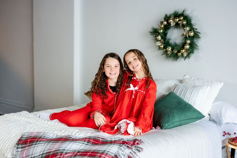 Two smiling girls in red Christmas pajamas sitting together on a white bed with wreath.