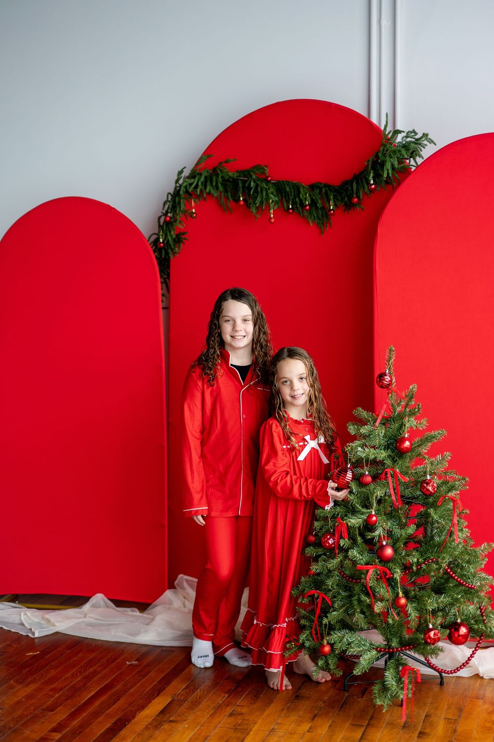 Two girls in red Christmas pajamas posing beside a decorated holiday tree with red backdrop.